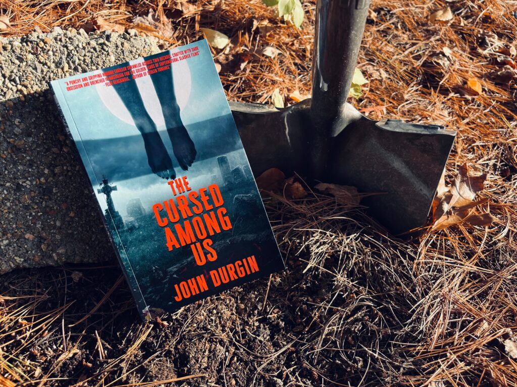 The Cursed Among Us by John Durgin book photo by Erica Robyn Reads - book next to a shovel