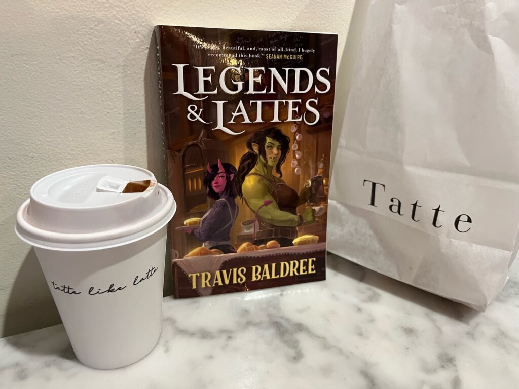 Legends & Lattes by Travis Baldree book photo by Erica Robyn Reads - Book sitting on a table in a cafe with a latte and bakery bag