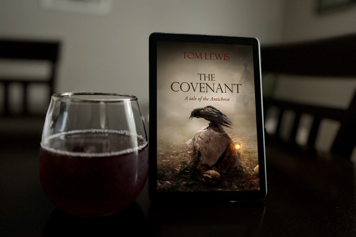 The Covenant: A Tale of the Antichrist by Tom Lewis | Book Review and Book Photo by Erica Robyn Reads - featuring a kindle ereader with the cover of the book displayed and a glass of red liquid. Book cover shows a raven sitting on a skull, lit by a candle.