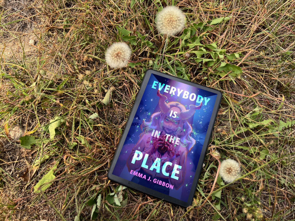 A tablet lies on dry grass and surrounded by dandelions in a natural outdoor setting. The screen displays the cover of the book Everybody Is in the Place by Emma J. Gibbon. The cover features a colorful, fantastical illustration of a female figure with glowing eyes and curved horns, holding something mysterious in her hands. Stars and swirling cosmic elements fill the background, giving the cover a magical and otherworldly feel. The title text is in bold, colorful letters across the top and middle, with the author's name at the bottom.