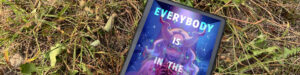 A tablet lies on dry grass and surrounded by dandelions in a natural outdoor setting. The screen displays the cover of the book Everybody Is in the Place by Emma J. Gibbon. The cover features a colorful, fantastical illustration of a female figure with glowing eyes and curved horns, holding something mysterious in her hands. Stars and swirling cosmic elements fill the background, giving the cover a magical and otherworldly feel. The title text is in bold, colorful letters across the top and middle, with the author's name at the bottom.
