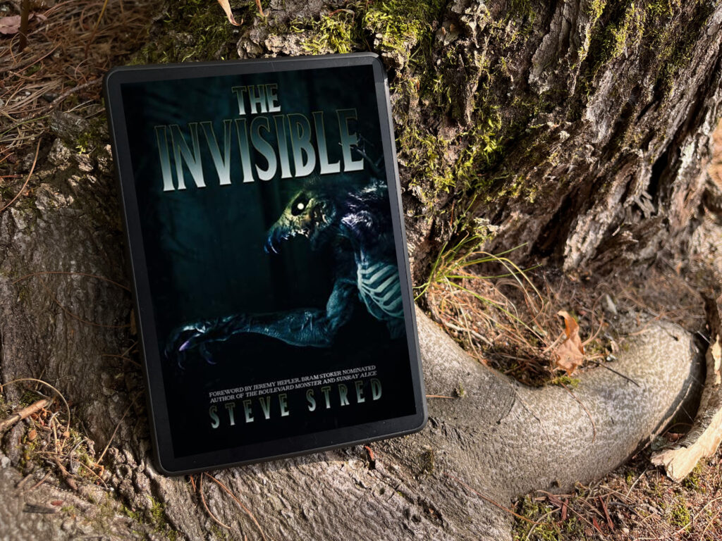 A tablet resting on a tree root in a forested area displays the cover of the book The Invisible by Steve Stred. The cover features a dark, eerie illustration of a monstrous creature with glowing eyes and sharp claws emerging from shadows. The title "The Invisible" is in large, bold, metallic lettering above the creature. Moss and fallen leaves surround the tree roots, enhancing the mysterious outdoor setting.